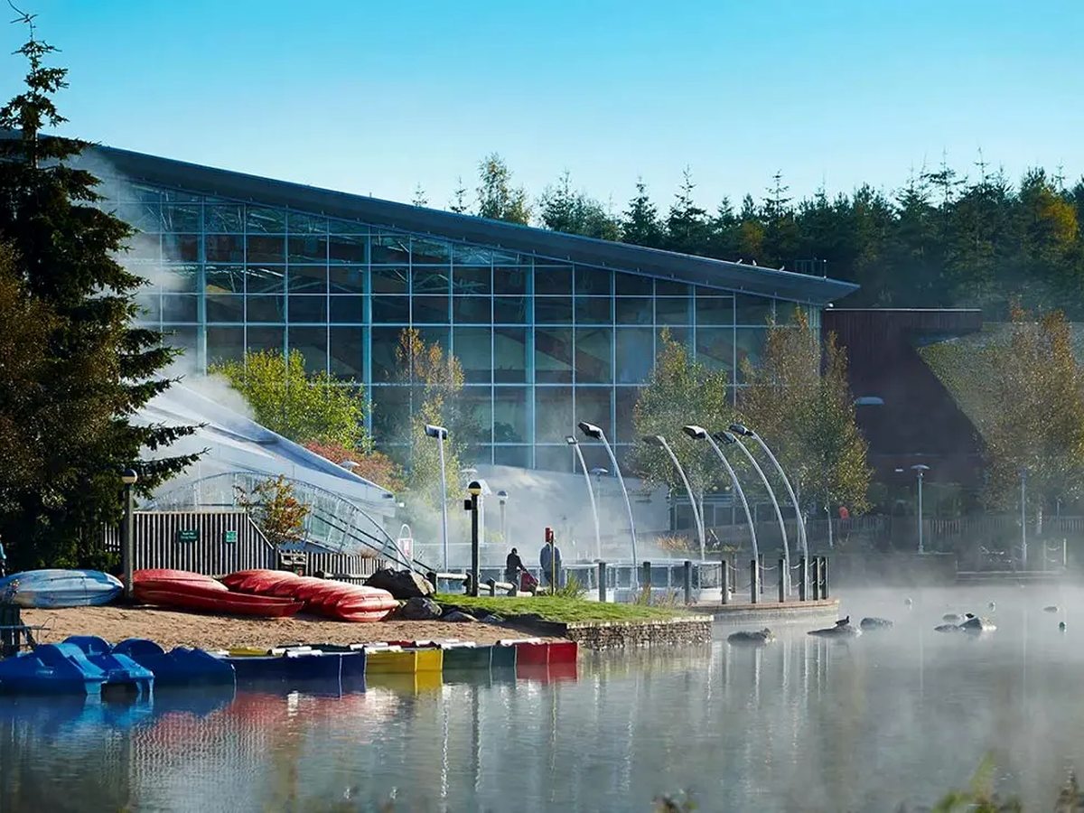 The long glass-fronted swimming complex above a sandy lake shore, with red canoes and blue pedalos drawn up on the sand and mist rising off the water.