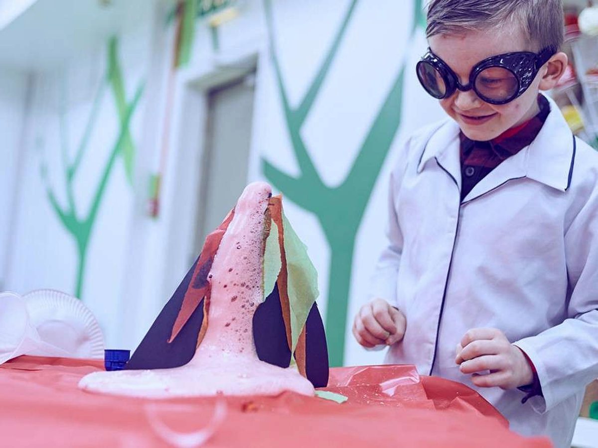 A young boy in a white lab coat and blacked-out goggles watching pink foam erupt from a paper volcano on a covered craft table.