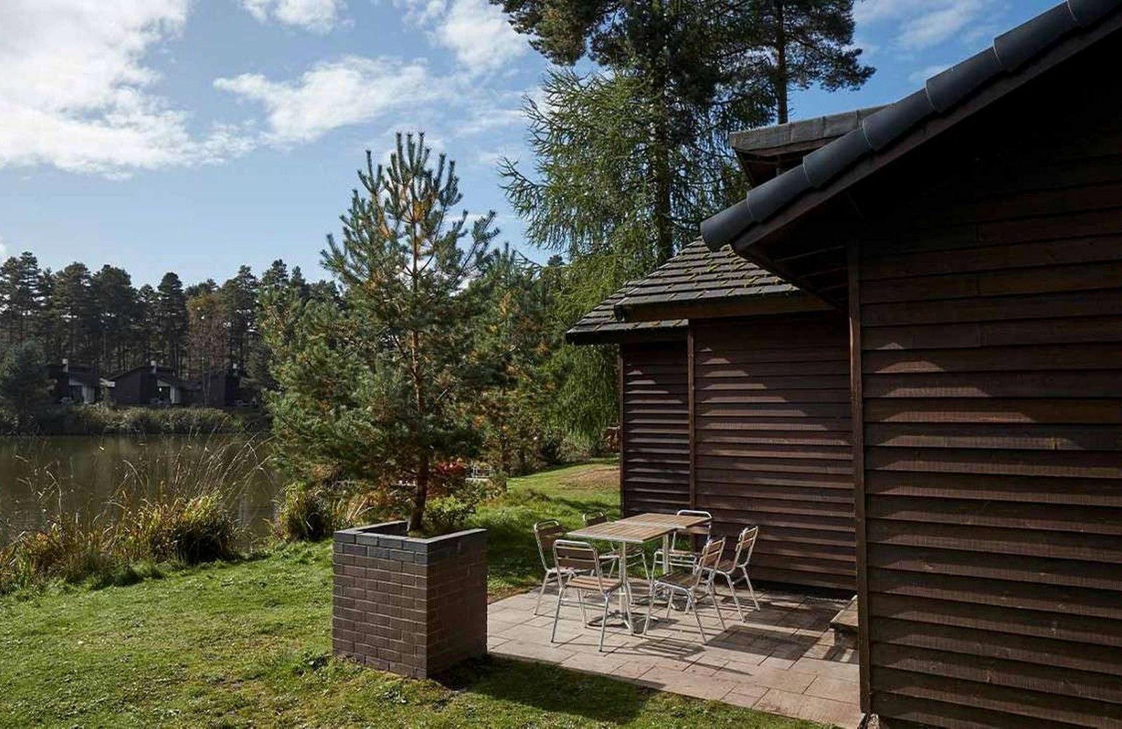 A dark timber lodge with a paved patio and an outdoor table and chairs on the edge of the lake at Whinfell Forest, with pines and more lodges across the water.
