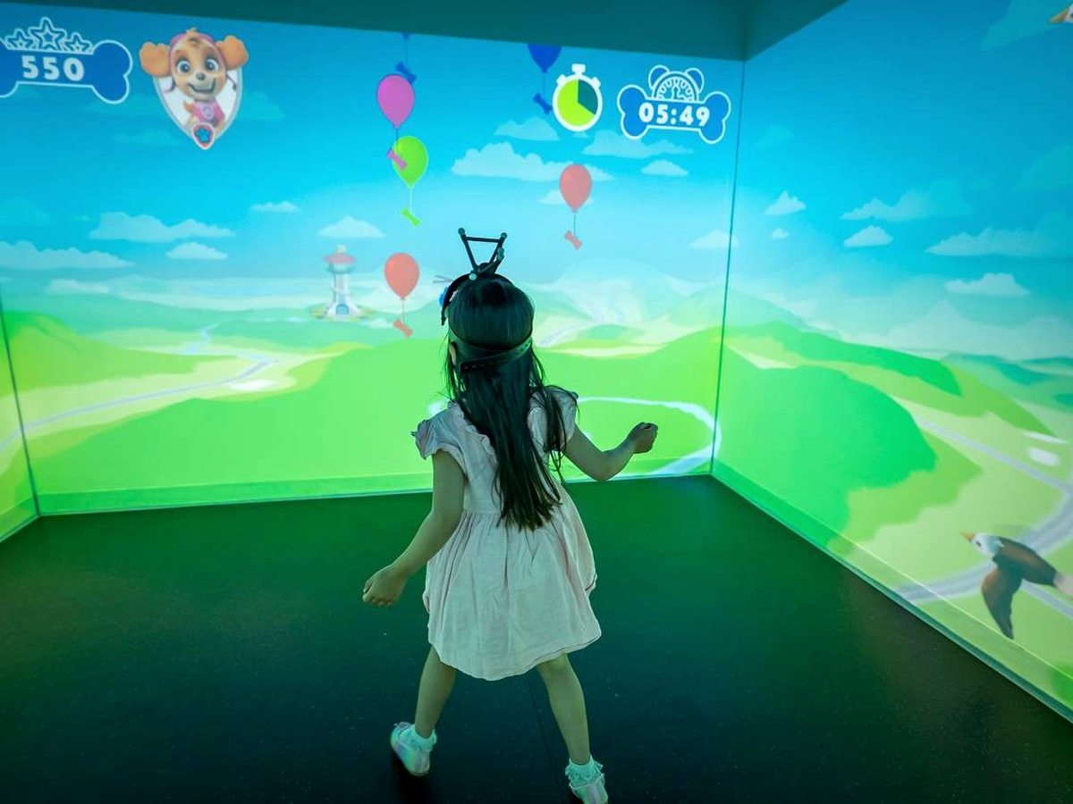 A young girl in a pale dress playing a cartoon balloon game projected across the walls and floor of a small room, with a score and a countdown timer shown above.