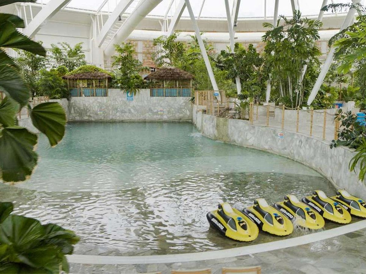 An indoor lagoon-shaped pool under a white domed glass roof, edged with stone walls, thatched shelters and dense tropical planting, with a row of yellow mini jet skis moored at the poolside.