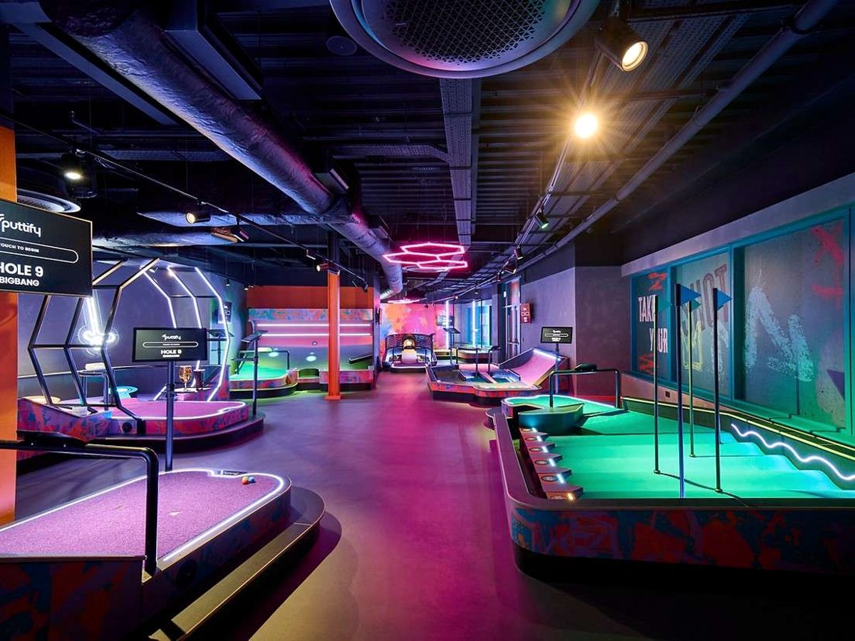 A neon-lit indoor mini-golf venue with green putting surfaces, patterned surrounds and digital hole screens under pink and teal lighting on an industrial ceiling.