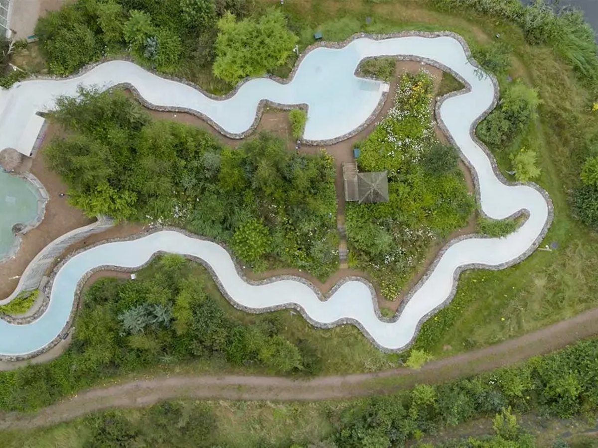 Aerial view of a wide white-bottomed water channel looping between stone edging, planted islands, gravel paths and a thatched shelter.