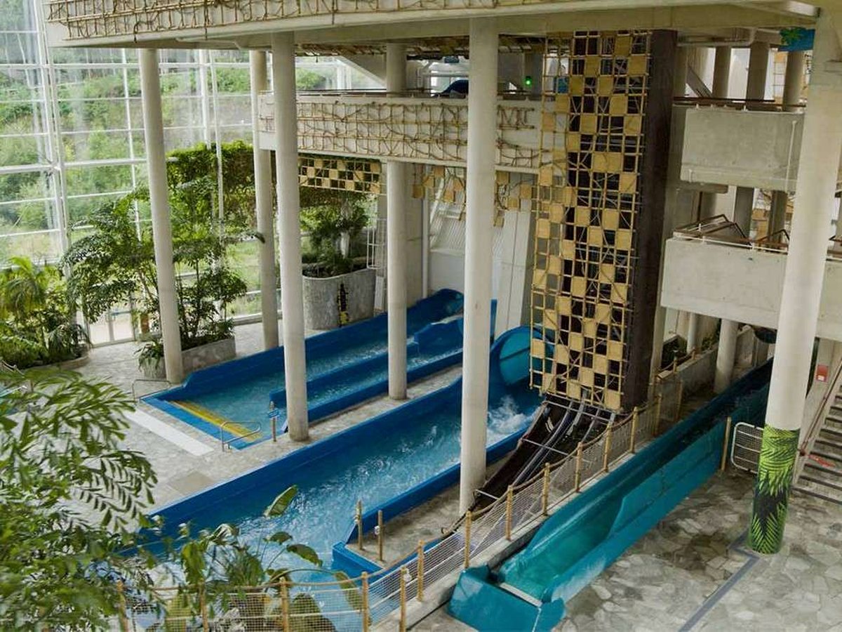 Blue and teal water flumes running side by side into a shallow indoor pool beneath concrete columns, rope bridges and a rattan-clad tower, with tall glazing and tropical planting to one side.