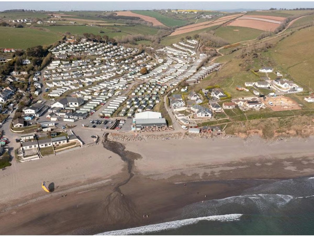 Aerial view of rows of static caravans and lodges covering a green hillside that runs down to a wide sandy beach and breaking surf.