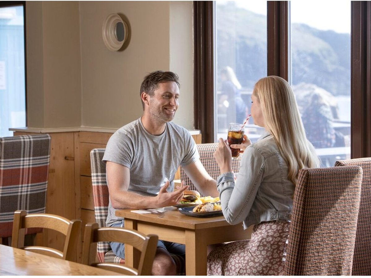 A couple eating burgers and chips at a wooden table beside tall windows, with tartan-upholstered chairs and a view out to the coast.