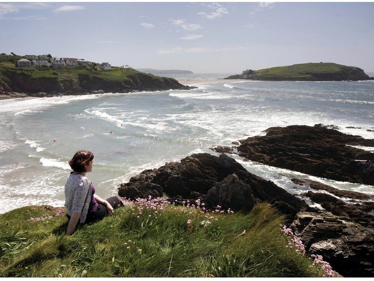 A woman sitting on a grassy, thrift-flowered clifftop looking across a surf-filled bay to a green tidal island topped with a building.