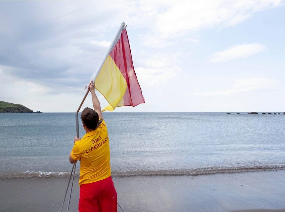 An RNLI lifeguard in red and yellow raising a red-and-yellow safety flag at the edge of the beach, with the calm bay and rocks behind.