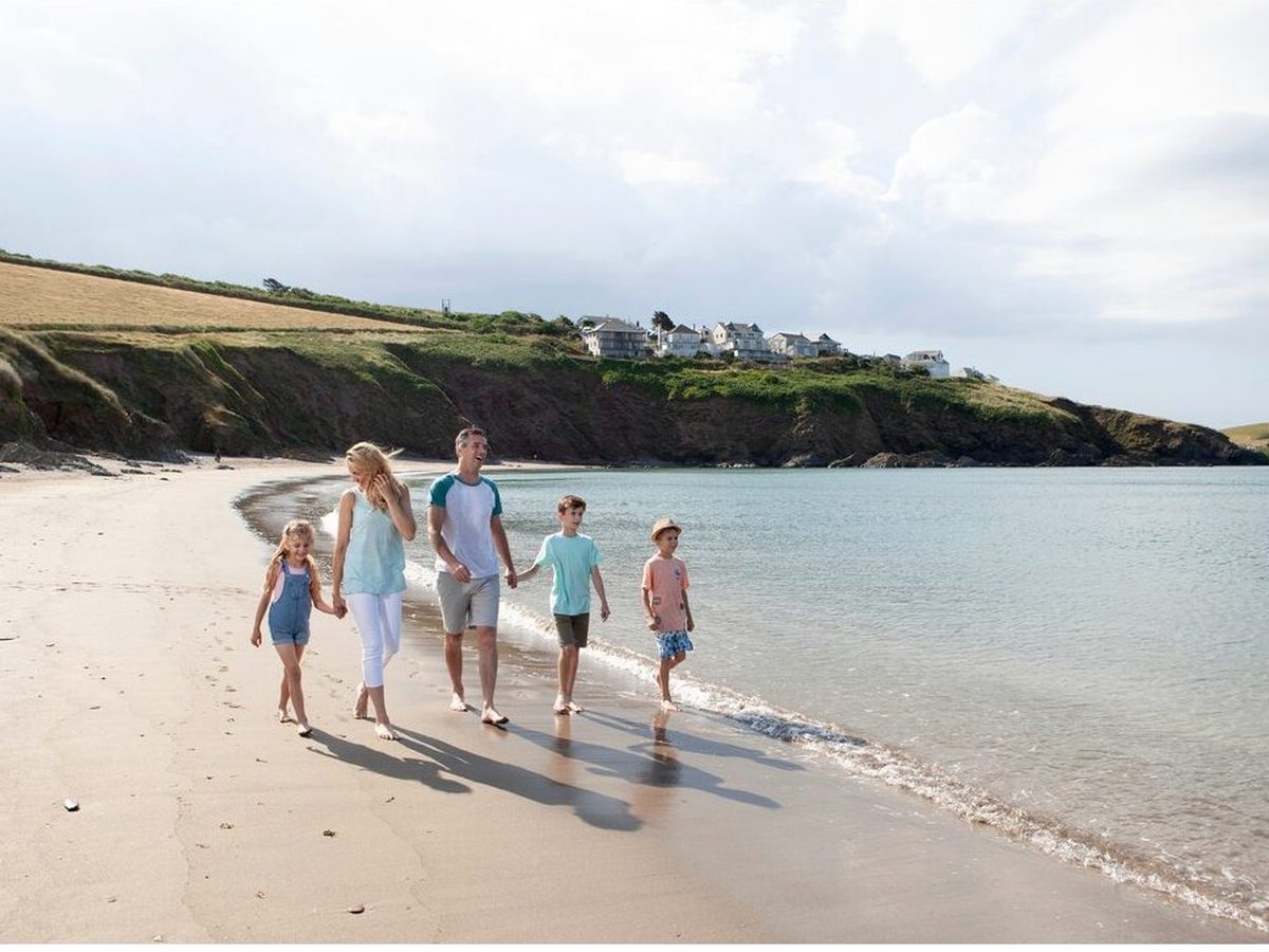 A family of five walking barefoot along the waterline of a sheltered sandy cove, with red cliffs and hillside houses behind.