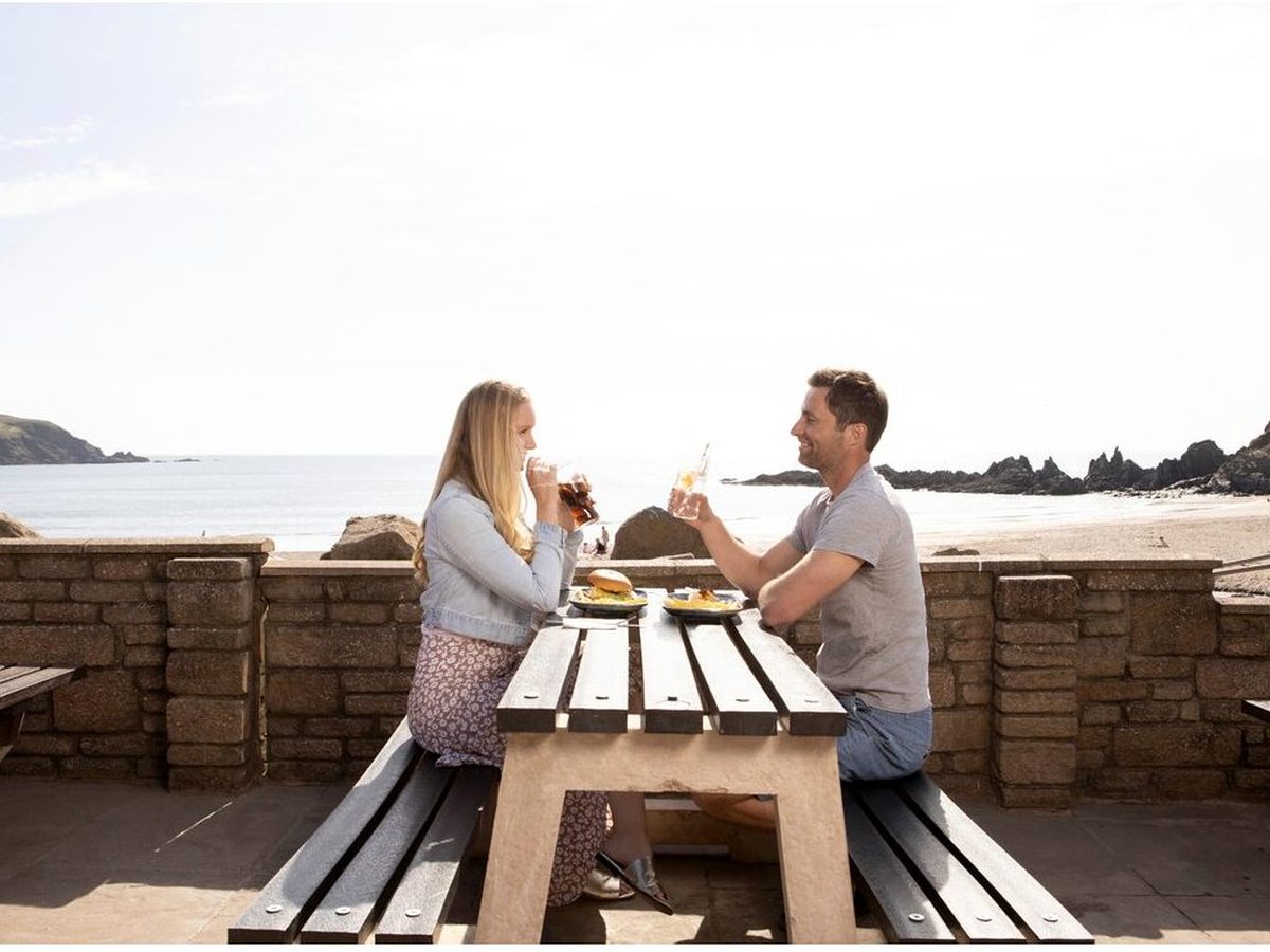 A couple raising drinks over lunch at a wooden picnic bench on a paved terrace, with a low stone wall and the sandy cove and rocks behind.