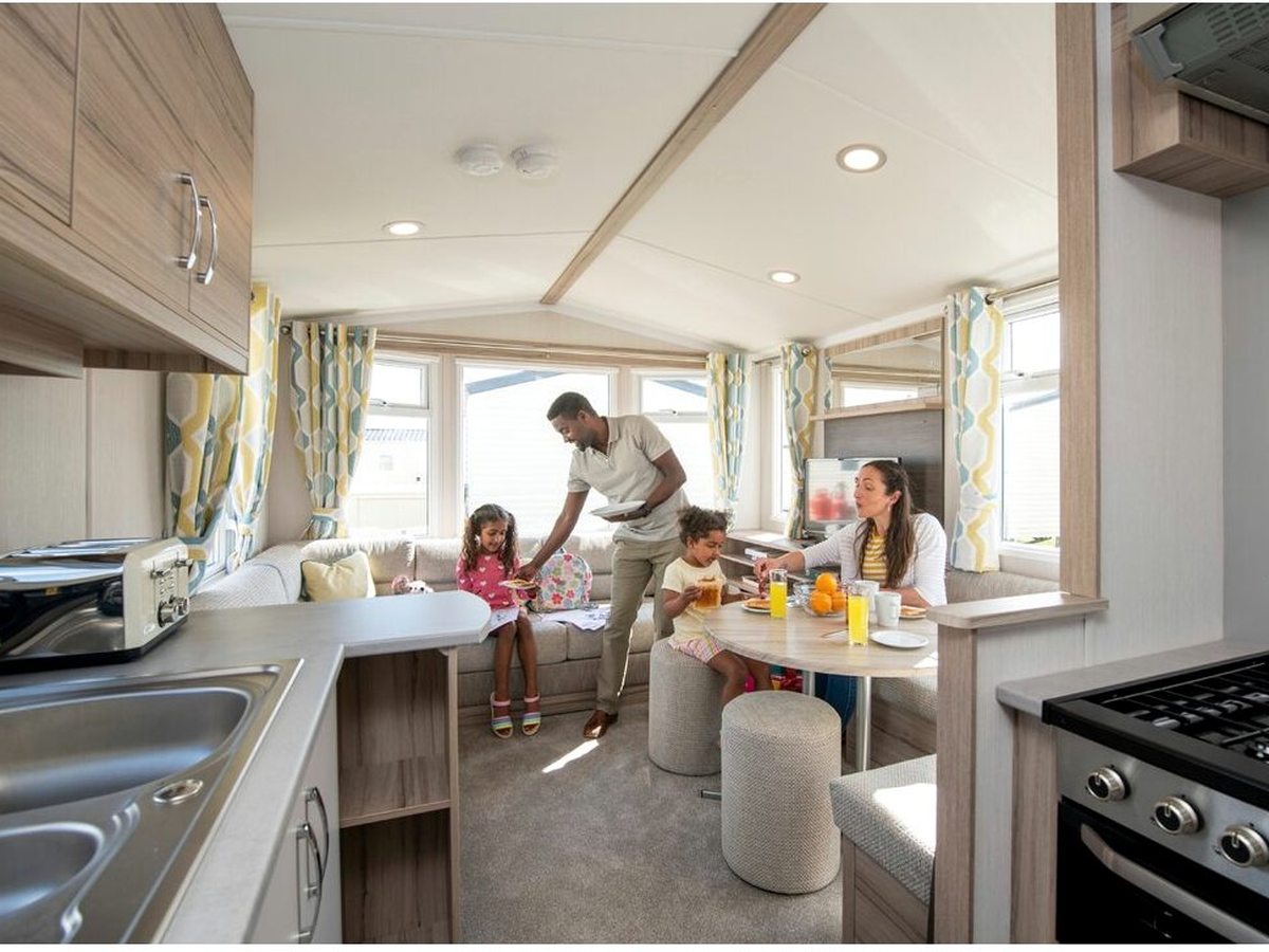 The open-plan interior of a static caravan, with a galley kitchen, gas hob and oven, a bay-window dinette and a family eating breakfast.