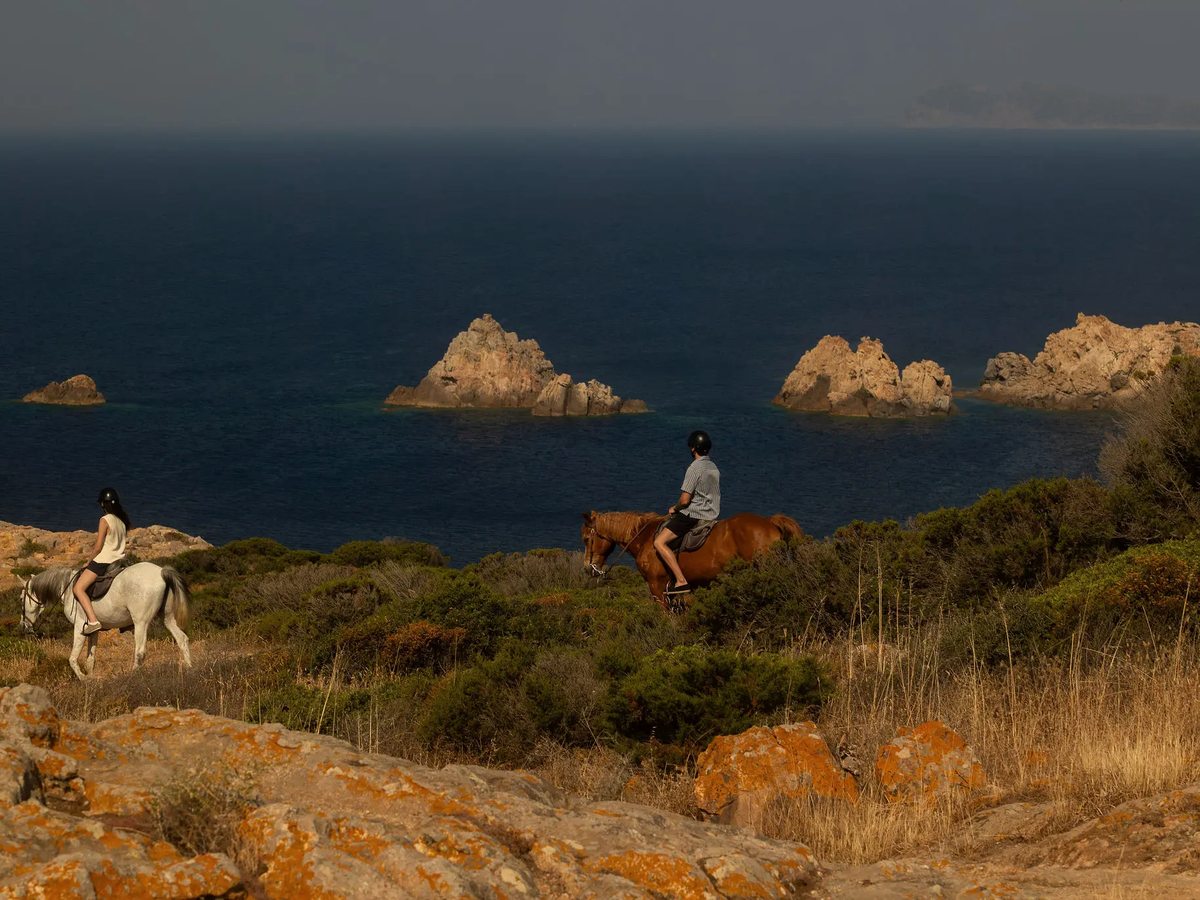 Two riders on horseback crossing a rocky, scrub-covered coastal headland above the open sea.