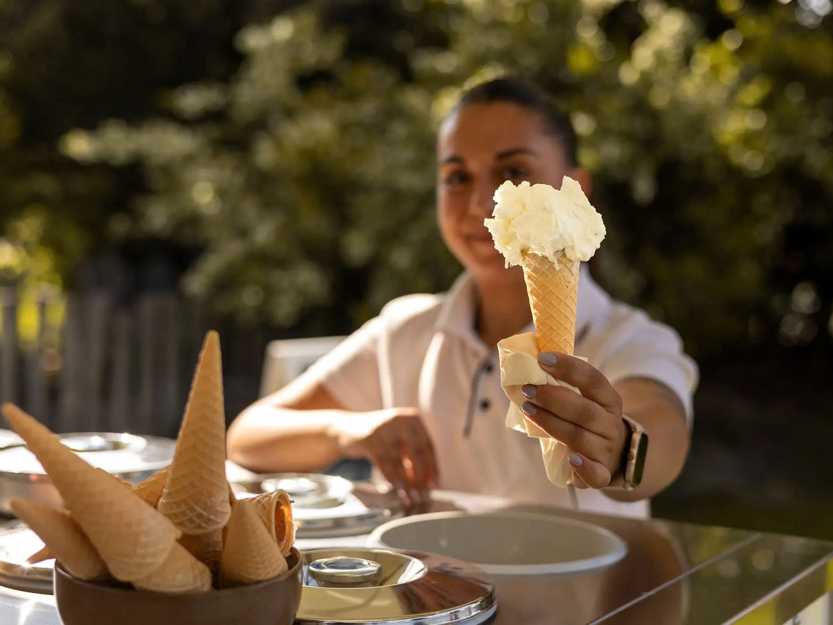 A member of staff in a white polo shirt holding out a cone of pale gelato across an outdoor gelato counter with a basket of empty waffle cones.