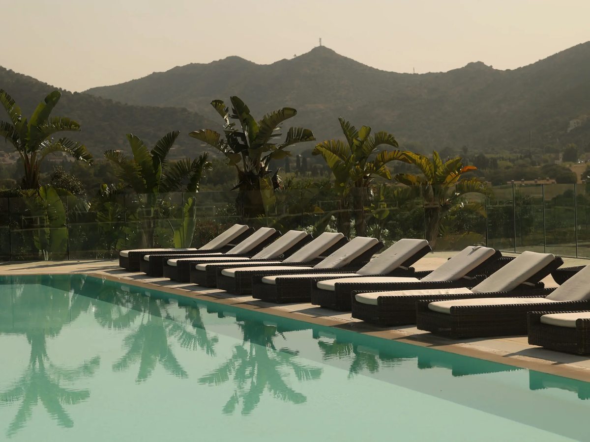 A long pool edged with a row of dark rattan sun loungers, tall palms and a glass balustrade, with scrubby green hills rising behind.