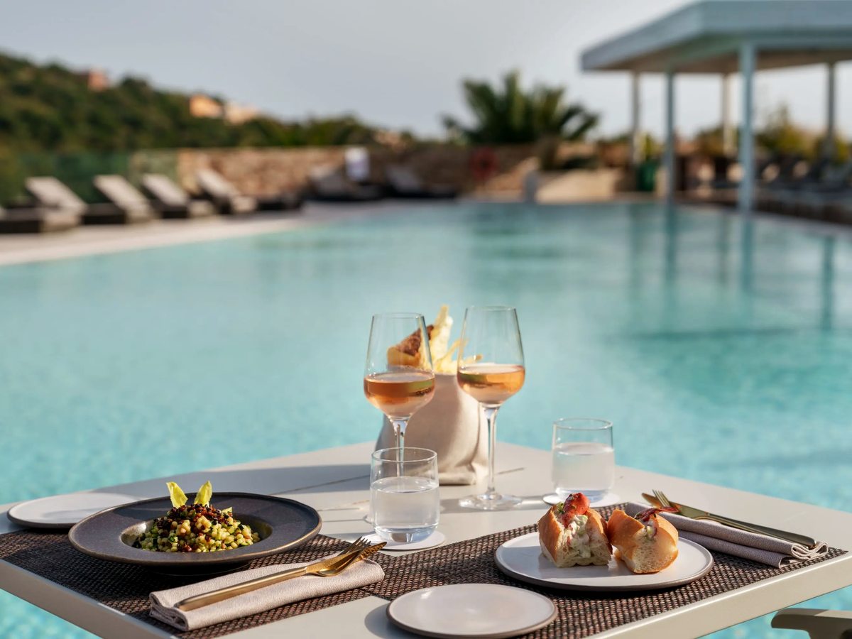 An outdoor table set with two glasses of rose wine, a salad and bread beside a bright turquoise pool lined with sun loungers and a poolside pavilion.
