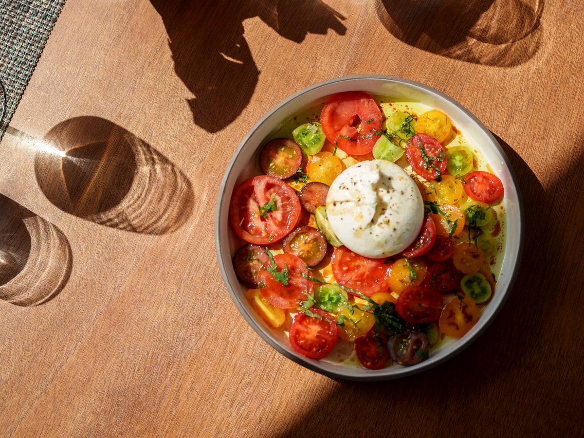 A bowl of sliced multicoloured tomatoes topped with a round of burrata cheese on a wooden table, seen from above with glasses alongside.