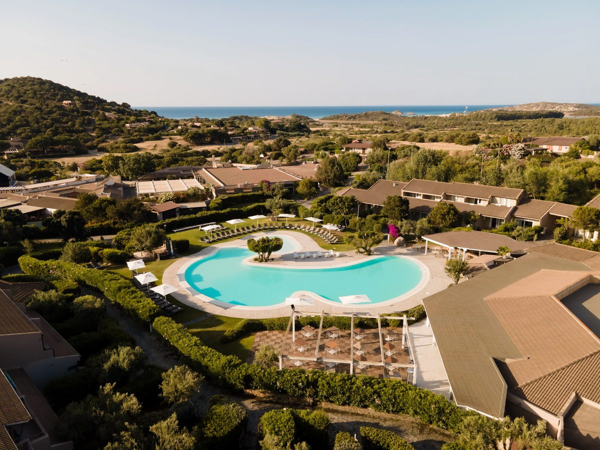 Aerial view of a large curved turquoise pool surrounded by paved terrace, lawns and low-rise cottage buildings, with the sea in the distance.