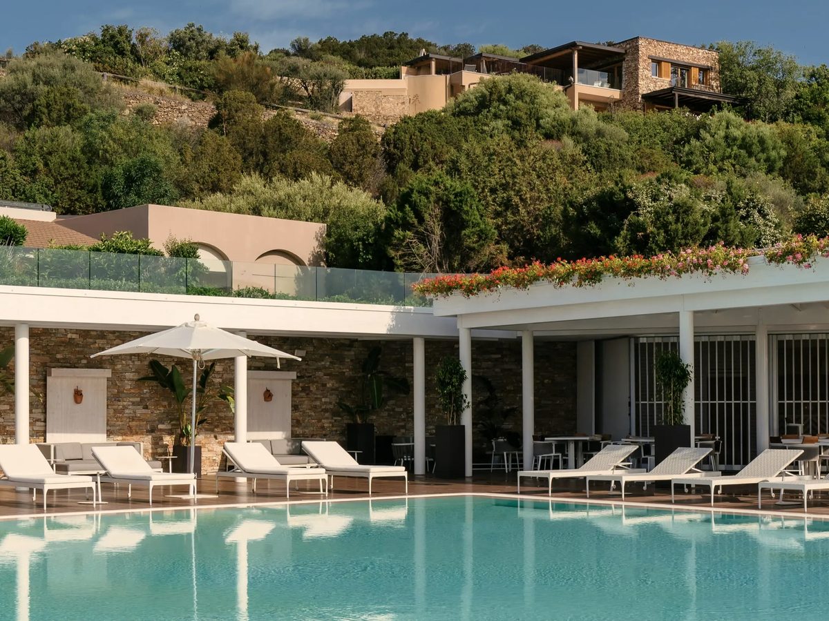 A rectangular pool with white sun loungers and a parasol along a stone-and-timber poolside terrace, with hillside villas and flowering planters above.