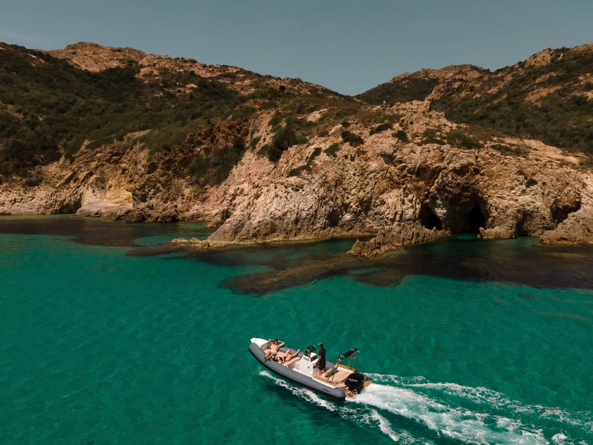 A small motorboat cruising through clear turquoise water past a rugged ochre-rock headland with sea caves under a bright sky.