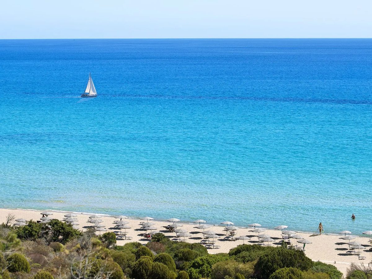 An elevated view over clear turquoise shallows deepening to deep blue, a small sailboat offshore and a strip of sandy beach with parasols below.