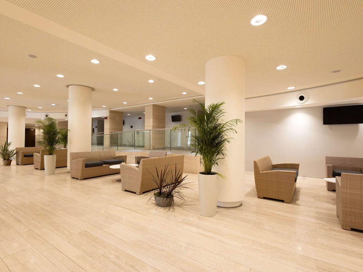 A bright open-plan hotel lobby lounge with cream wicker sofas and armchairs, tall round columns and potted palms on a pale tiled floor.