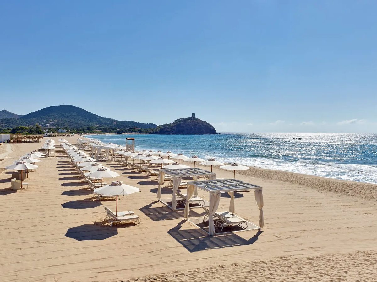 A long flat sandy beach lined with rows of white parasols and shaded beds, the sea to the right and the Chia Tower on a headland in the distance under a clear blue sky.