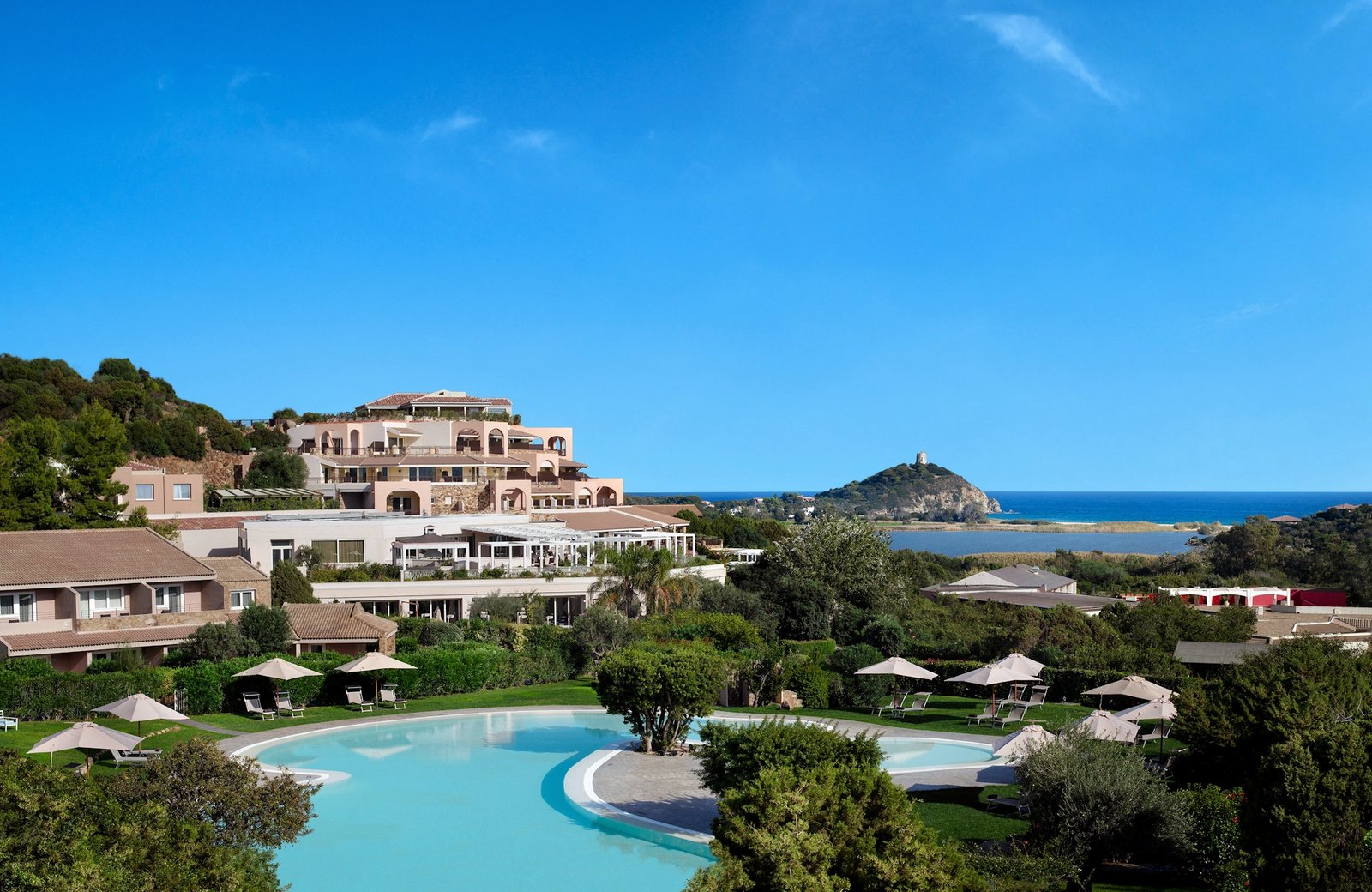 Chia Laguna above the Chia coast in southern Sardinia: a terraced hotel and a curved turquoise pool, with the Chia Tower on the headland and the lagoon and sea beyond.