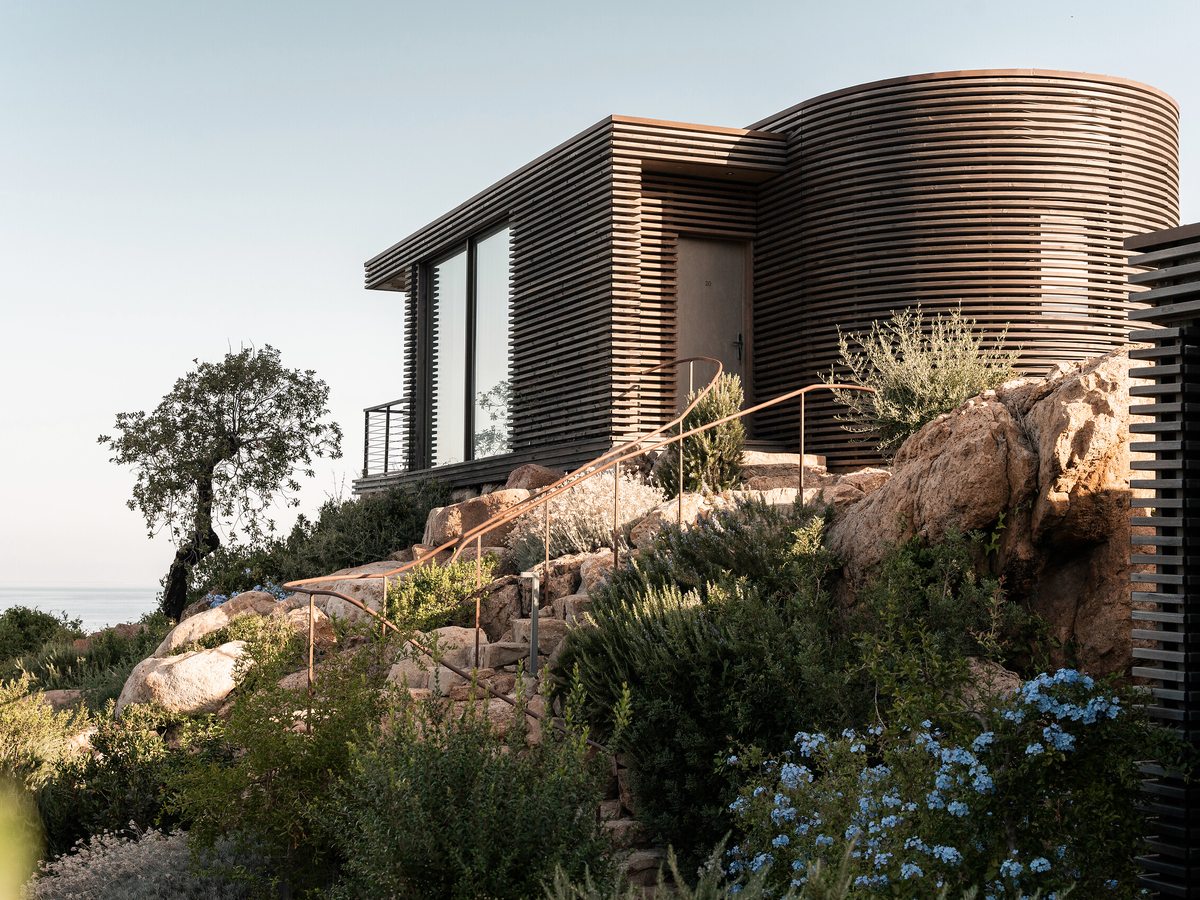 A timber-slatted resort building with a covered balcony on a rocky, planted slope with a partial sea view in warm light.