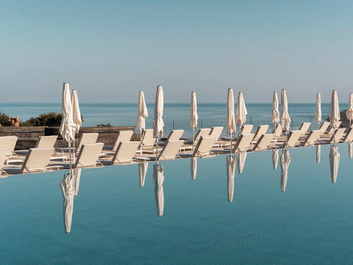 A row of white sun loungers with closed white parasols along a pool edge that meets the flat blue sea and sky.