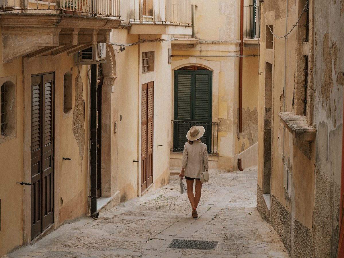 A narrow sunlit lane of cream stone buildings with shutters and balconies in Cefalù's historic centre, a woman in a sun hat walking away.