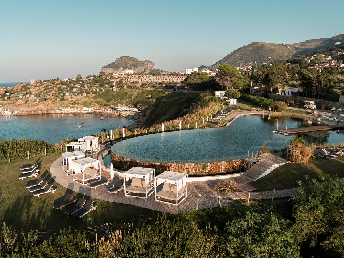 A large curved pool with white canopied cabana daybeds on a timber deck, overlooking Cefalù Bay with hills and a crag beyond.