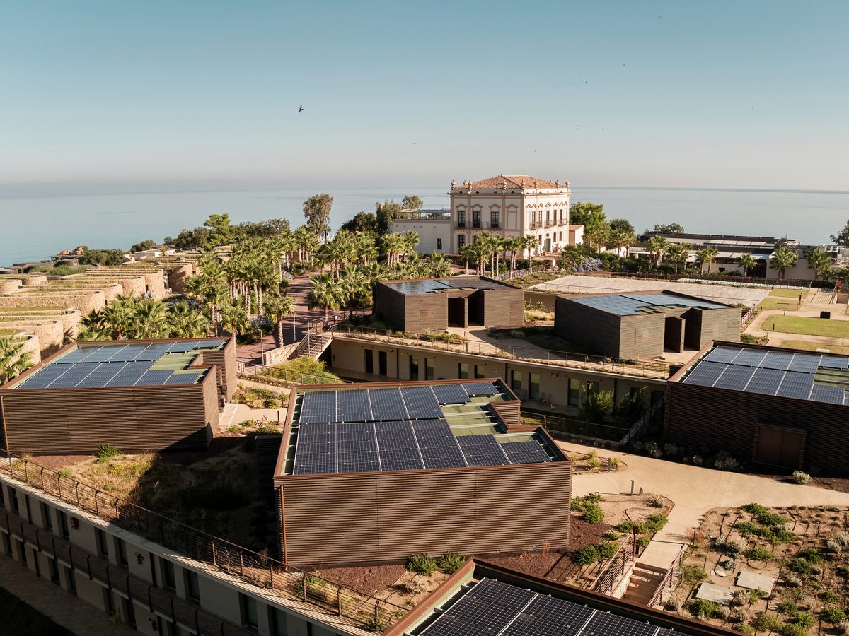 Aerial view of low timber-clad resort buildings with solar-panelled and planted roofs, terraced across a headland above the sea.