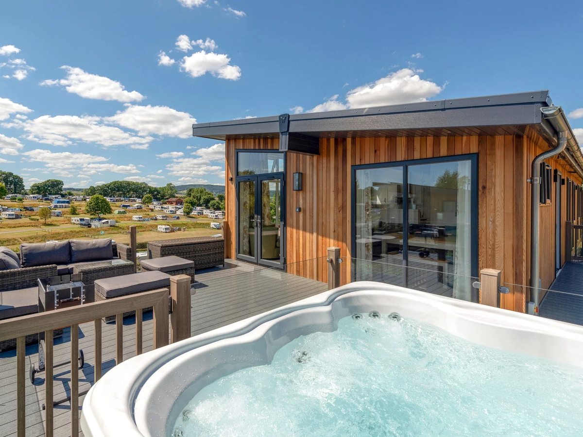 A timber-clad lodge with full-height glass doors and a decked terrace holding a private hot tub and rattan sofas, looking out over the park under a blue sky.