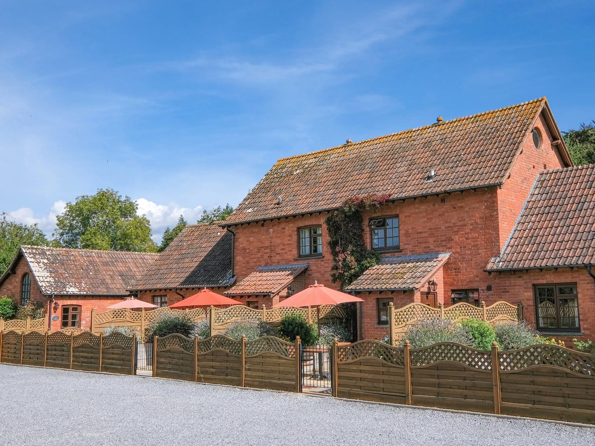 A row of red-brick barn-conversion holiday cottages with tiled roofs, behind a low trellis fence with lavender beds and coral parasols under a blue sky.