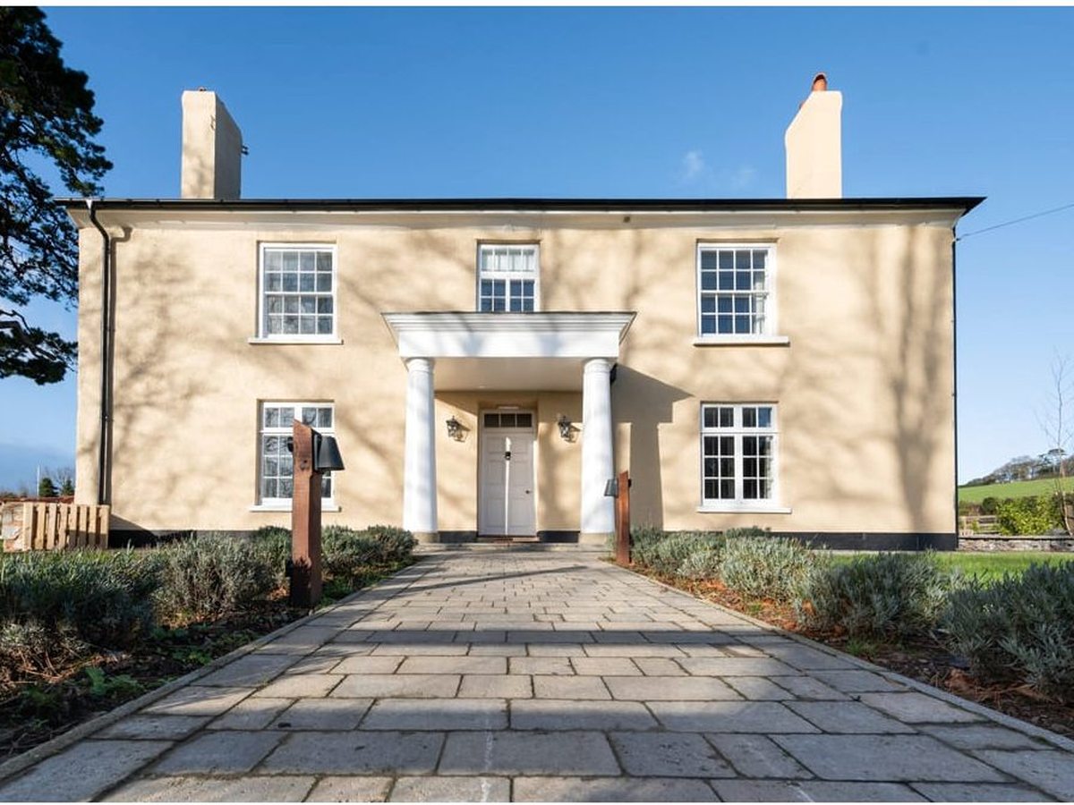 A cream two-storey Georgian farmhouse with a columned porch, sash windows and chimneys, a paved path leading to the front door across a lawn under a blue sky.