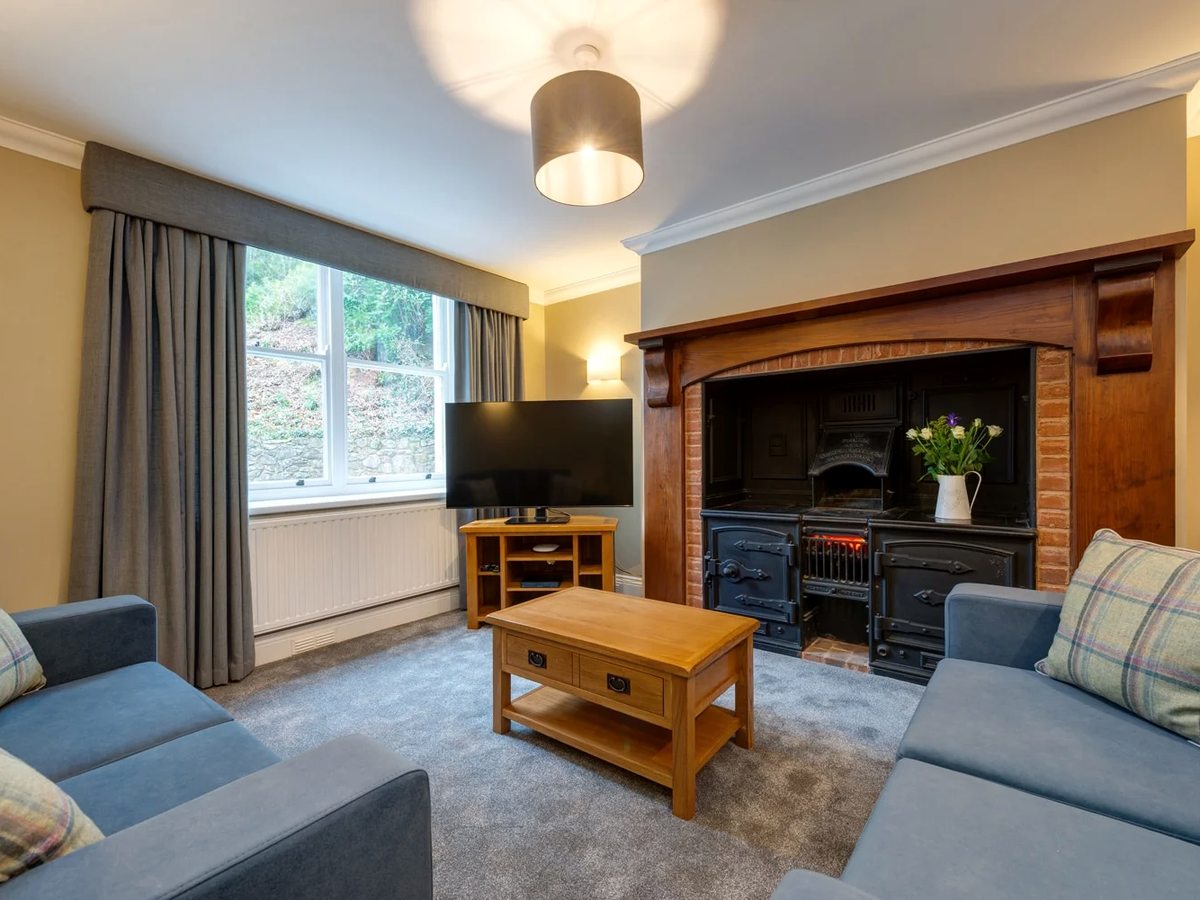 A holiday cottage living room with blue sofas, a wood-effect electric stove in a brick fireplace and a coffee table on a patterned carpet.