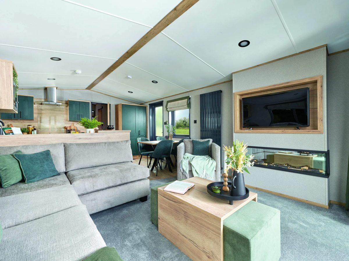 A modern static caravan living area with a grey corner sofa, green cushions, a wall-mounted TV and an open kitchen and dining space beyond.