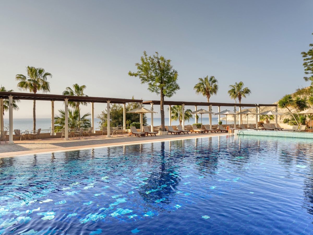 A long blue-tiled infinity pool edged by a pergola and sun loungers, with palms and the sea beyond.