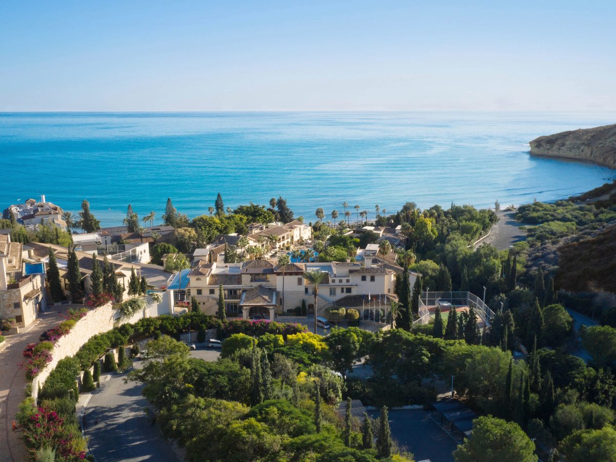 An aerial view of the honey-coloured resort buildings among gardens and cypress trees, above the curving bay and headland.