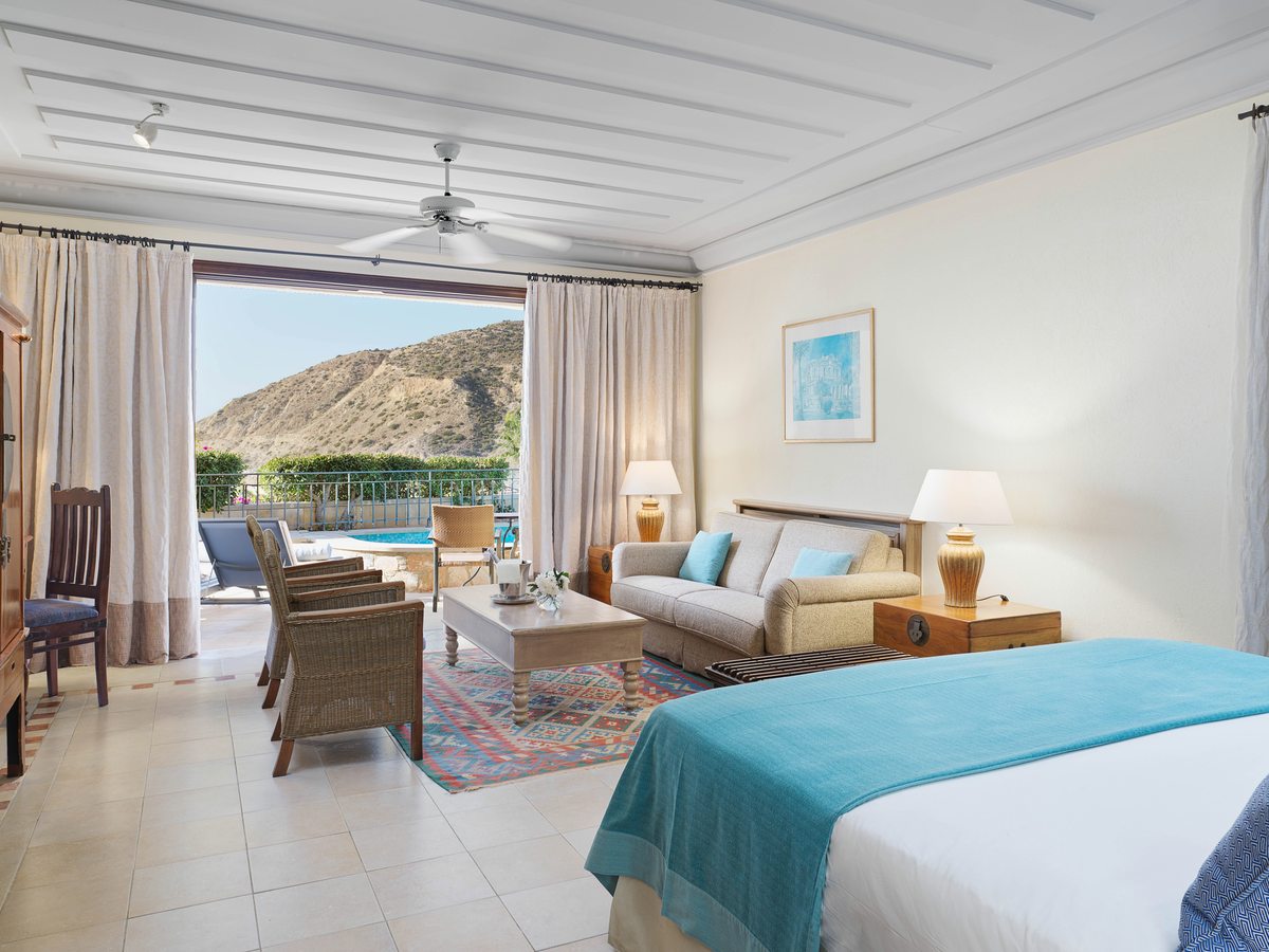 A suite bedroom with a double bed and sitting area, full-height curtains and doors opening to a terrace with a cliff view.