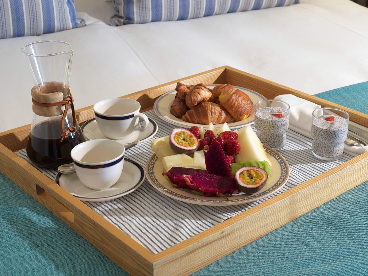A wooden breakfast tray with a cafetiere, coffee cups, pastries, cheese and fruit on a bed.