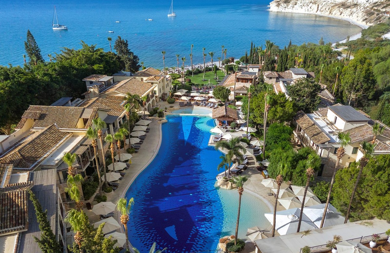 An aerial view of Columbia Beach Resort's large lagoon-style pool ringed by terracotta-roofed suites and palms, with Pissouri Bay, sailboats and white cliffs beyond.