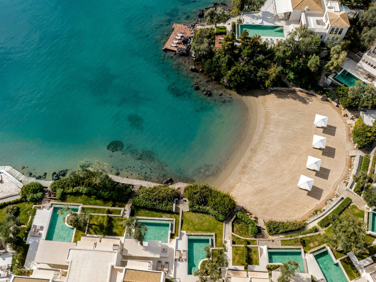 Aerial view of a small sandy cove with white parasols on the sand, clear turquoise water and beachfront villas with private pools stepping down to the shore above.