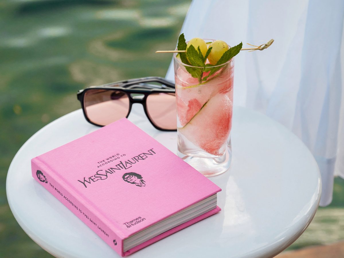 A tall iced fruit cooler garnished with mint and grapes on a small white table beside the water, with a book and sunglasses.