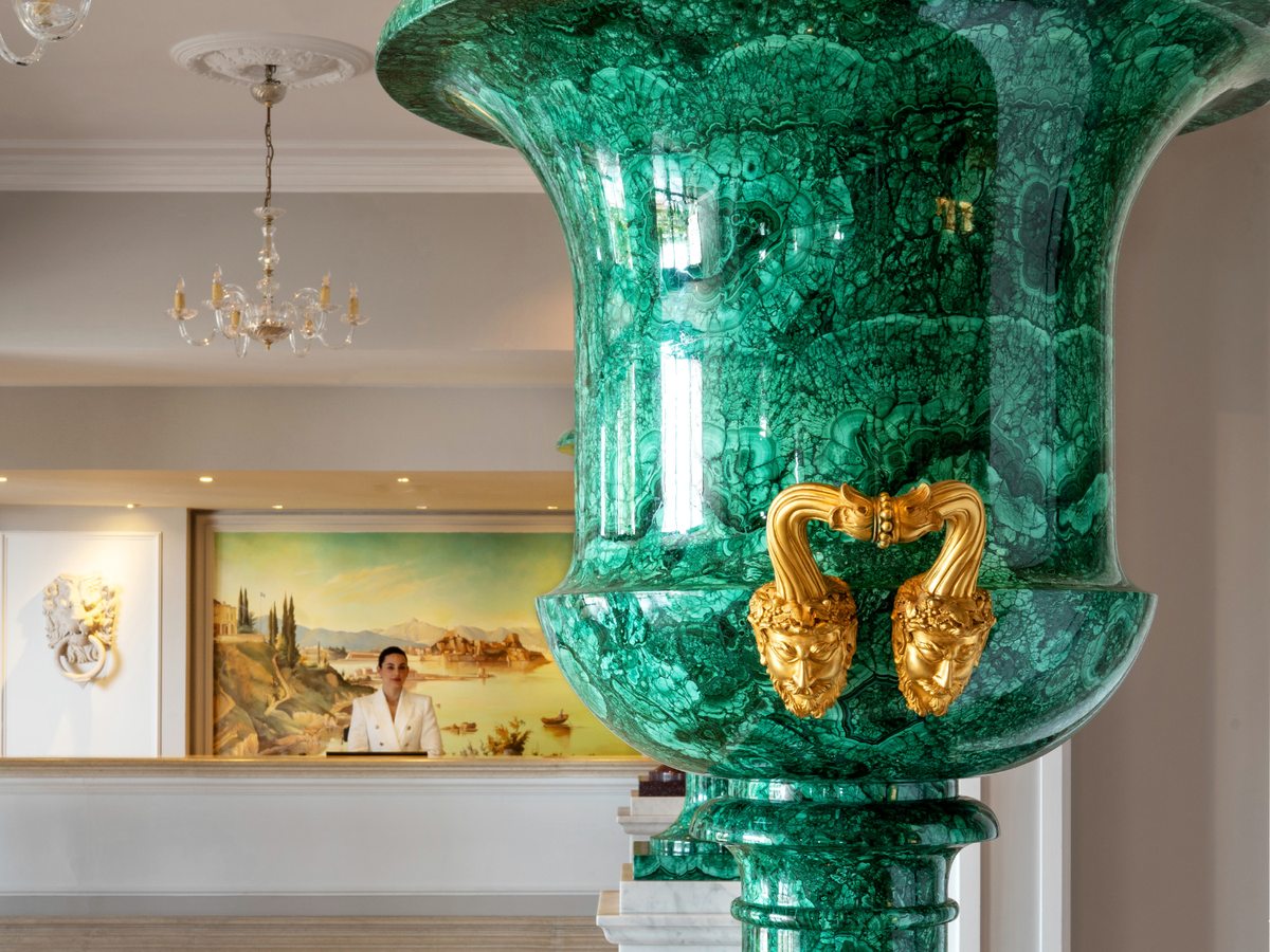 Hotel reception desk with a large green malachite-style urn with gilded handles, a chandelier, staff behind the counter and a painted landscape mural on the wall.