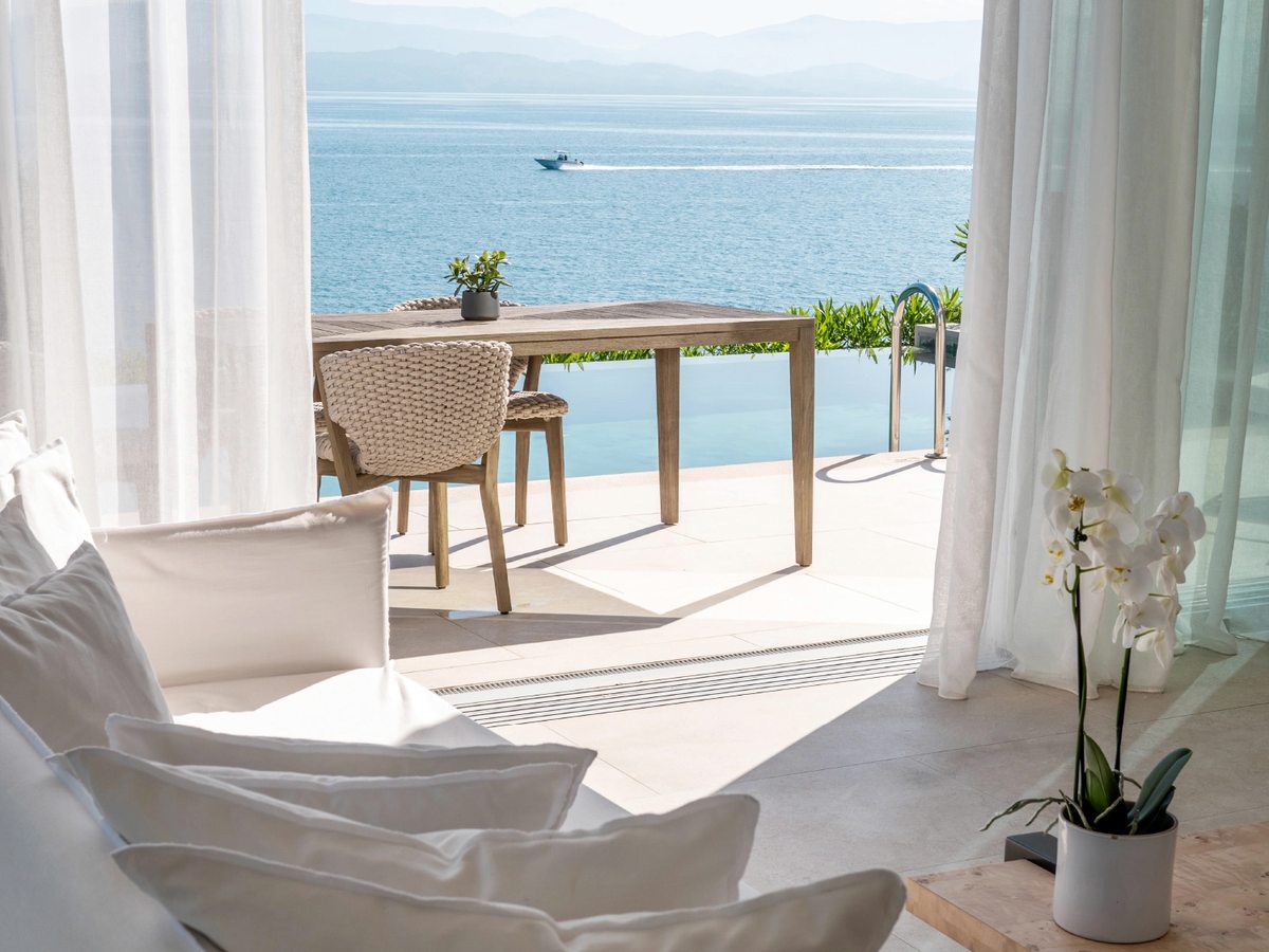 Suite lounge with white seating and sheer curtains opening to a private balcony with a table and chairs, a potted orchid and a panoramic view over the sea.