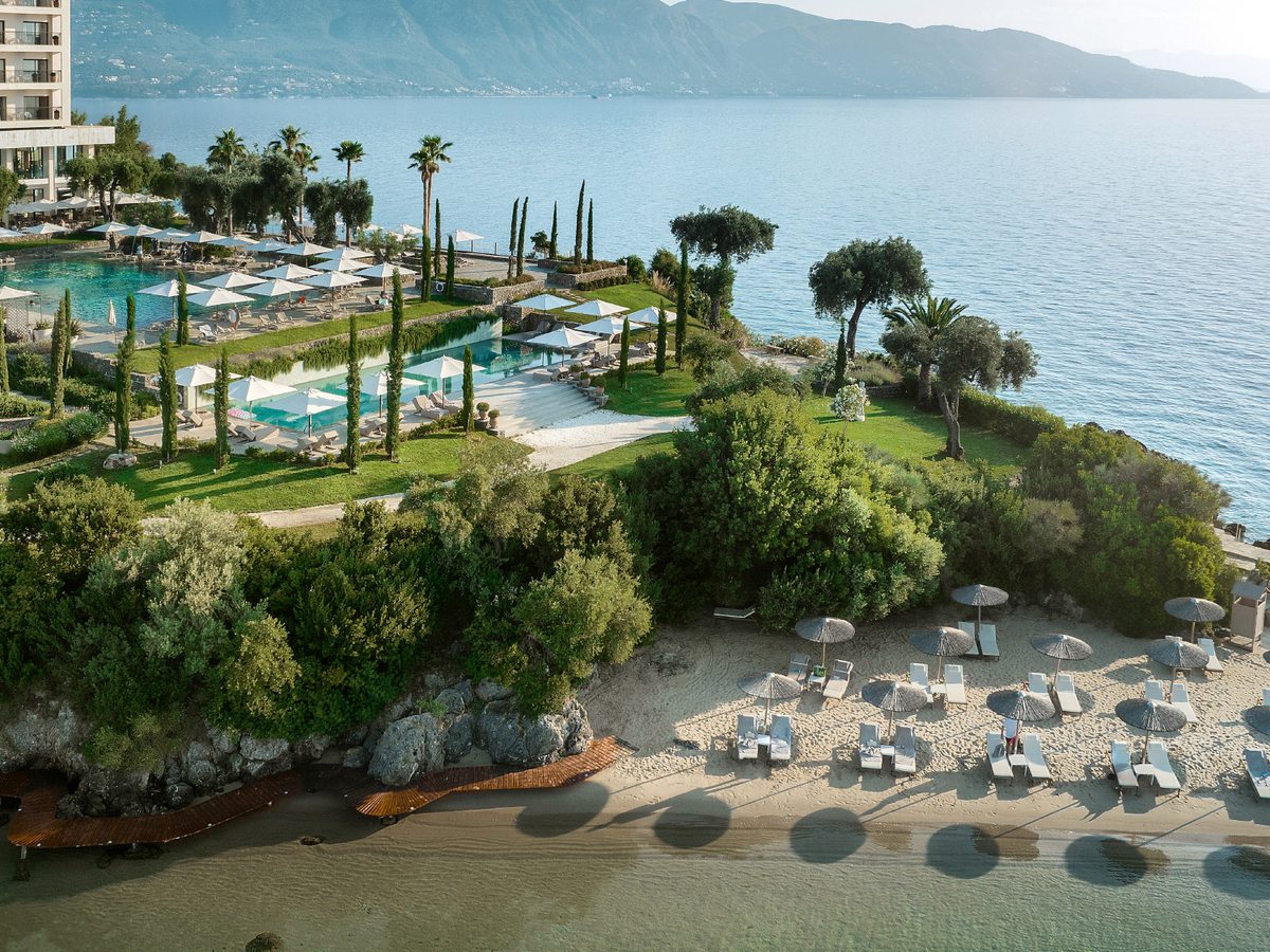 A sandy beach with grey parasols and sun loungers below the resort, terraced pools and gardens rising behind it and mountains across the bay.