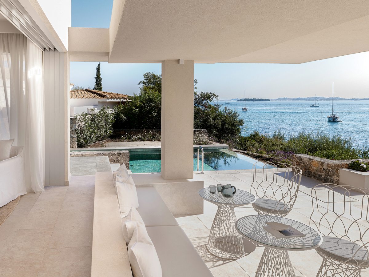 Covered villa terrace with white sofas and outdoor seating beside a private pool, looking over the sea to moored boats.