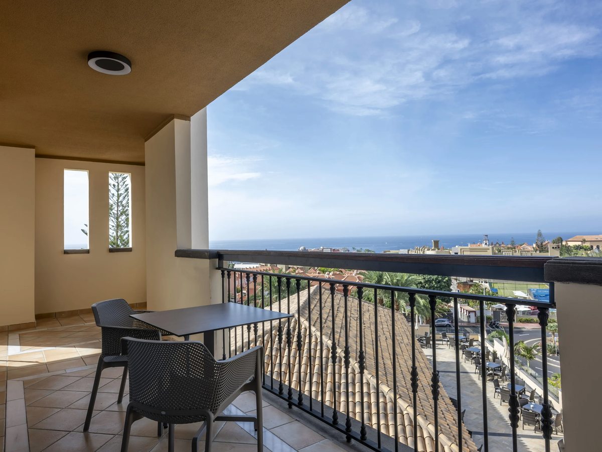 A covered room balcony with a table, two chairs and a black iron railing, opening to a wide view over the coast to the sea.