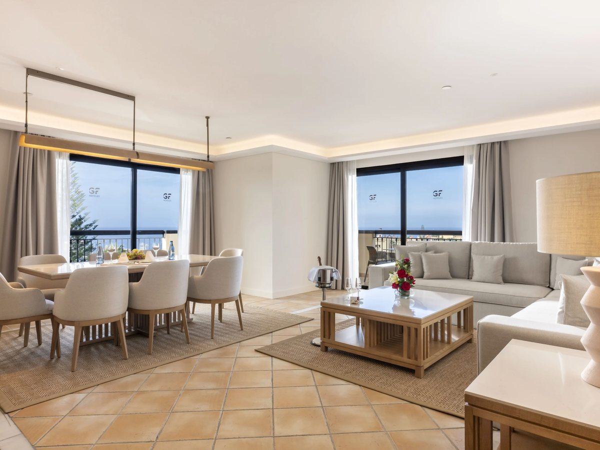 A suite's open-plan living and dining space with sofas, a dining table and a wall of balcony windows looking to the sea.